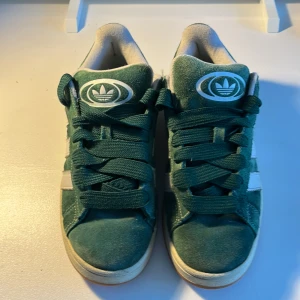 Adidas Campus gröna sneakers mocka - Gröna Adidas Campus sneakers i mocka med klassiska vita ränder på sidorna och gummisula. Skorna har snörning och rund tå, samt Adidas logga på plösen. Perfekta för dig som gillar retrostil och streetwear.