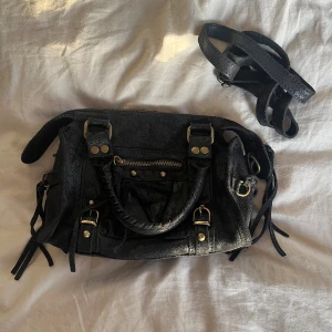 Svart handväska/axelväska kostar 900kr ny - Cool svart axelväska i skinn med guldiga metalldetaljer, dragkedja framtill och flätat handtag. Väskan har flera dekorativa remmar och nitar samt en avtagbar axelrem. Perfekt storlek för det viktigaste och ger en edgy vibe till din outfit.