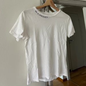 Vit t-shirt med text vid halsen - Vit t-shirt i bomull med broderad text 'BE A DREAMER' vid halsringningen. Klassisk passform perfekt för en clean och enkel stil.