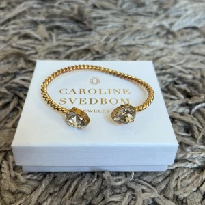 Caroline Svedbom armband - Snyggt armband från Caroline Svedbom i guldtonad metall med tvinnad design. Armbandet är öppet och pryds av två stora, gnistrande kristaller i båda ändar som ger en lyxig känsla. Perfekt statement piece för att lyfta din look.