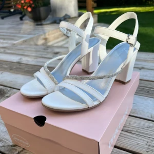 Vita sandaler med klack från Graceland - Stilrena vita sandaler från Graceland med bred klack och öppen tå. Remmarna har snygga detaljer med glittrande stenar och spänne runt ankeln. Använda på ett bröllop 
