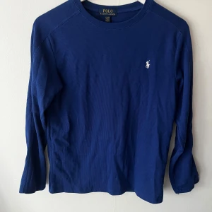 Blå långärmad tröja Polo Ralph Lauren - Snygg blå långärmad tröja från Polo Ralph Lauren med klassisk vit logga på bröstet. Tröjan är ribbad och har en enkel, stilren design som passar perfekt till jeans eller chinos. Tillverkad i mjuk bomull för skön känsla hela dagen.