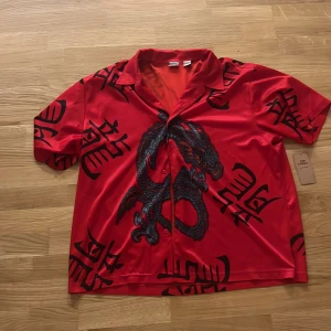 Röd skjorta med draktryck - Snygg röd skjorta med svart drakmotiv och asiatiska tecken. Skjortan har korta ärmar, knappar framtill och en loose fit. Materialet är glansigt och känns mjukt mot huden, perfekt för dig som vill sticka ut med en unik stil.