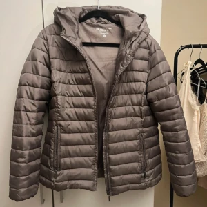 Brun pufferjacka från Champion - (Mindre i storlek) Säljer en brun pufferjacka från Champion med huva och dragkedja framtill. Jackan har quiltad design, två fickor med dragkedja och en snygg, stilren look. Perfekt för kyliga dagar och enkel att matcha med olika outfits.