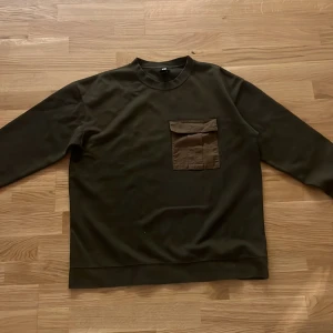Olivgrön sweatshirt från H&M - Olivgrön sweatshirt från H&M med en stor bröstficka i matchande färg. Tröjan har rund halsringning och långa ärmar. Perfekt för en chill och avslappnad stil.