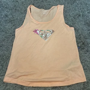 Persikofärgad linnetopp från Roxy - Snygg persikofärgad linnetopp från Roxy med ett stort hjärtformat logotryck i mitten, dekorerat med blommigt mönster och en rosa blomma. Tunt och mjukt material, perfekt för varma dagar. Ärmlös och med rund halsringning.