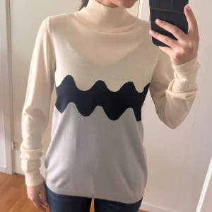 Beige och grå polotröja med vågmönster - Snygg polotröja i beige och ljusgrått med ett svart vågigt mönster över bröstet. Tröjan har lång ärm, ribbade muddar och hög krage. Perfekt för dig som gillar stilrena plagg med en twist. Materialet känns mjukt och lätt, passar till jeans eller kjol. Endast använd 2 gånger, väldigt bra skick! 