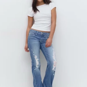 Blå slitna bootcut jeans Gina Tricot - Snygga blå bootcut jeans från Gina Tricot med slitna detaljer framtill och coola bakfickor med knapp. Jeansen har låg midja och klassisk femficksdesign. Perfekta för en avslappnad och trendig look.