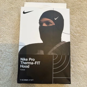 Nike Pro Therma-FIT Hood svart - Nike Pro Therma-FIT Hood i svart med vit Nike-logga. Snygg och tight balaclava som täcker hela huvudet och halsen, perfekt för träning och fotboll. Tillverkad i mjukt och stretchigt syntetmaterial som håller dig varm och bekväm även när det är kallt ute.