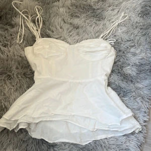 Vit korsett-topp från Zara XS - Supersöt vit korsett-topp från Zara i storlek XS. Toppen har tunna axelband som knyts, markerade kupor och lätt utsvängd nederdel. Perfekt för att skapa en trendig och feminin look. Gjord i bomull för en skön känsla.