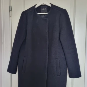 Svart kappa från Monki, storlek S - Stilren svart kappa från Monki i storlek S. Kappan har minimalistisk design med raka linjer, diskreta fickor och rund halsringning. Materialet är en mix av ull, polyester och polyamid, vilket gör den både varm och mjuk. Perfekt för dig som gillar en clean look.