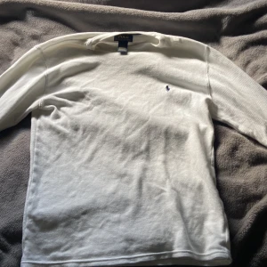 Vit långärmad tröja från Polo Ralph Lauren - Klassisk vit långärmad tröja från Polo Ralph Lauren i 100% bomull. Tröjan har ett diskret rutmönster i strukturen och den ikoniska broderade loggan på bröstet. Perfekt basic med rund hals och ribbade muddar.