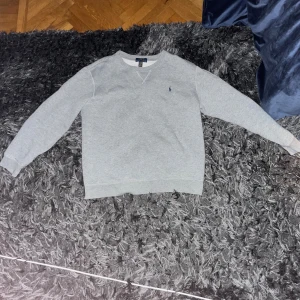 Grå sweatshirt från Polo Ralph Lauren - Klassisk grå sweatshirt från Polo Ralph Lauren med broderad marinblå logga på bröstet. Storlek M stor S Tröjan har rund halsringning, ribbade muddar och är tillverkad i mjuk bomullsmix. Perfekt för en chill och stilren look.