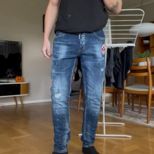 Dsquared2 Slim Skater Jeans (1964 fit) - Säljer ett par blå jeans från Dsquared2 med coola slitningar och en rödvit patch med lönnlöv på ena benet. Tyget är bekvämt stretchigt och passformen är tajt. Paint-faded/distressed finish. Riktigt snygga, går ej att hitta någonstans! Är 177cm 70kg. Mina ben är något längre så passar för män 175-185.