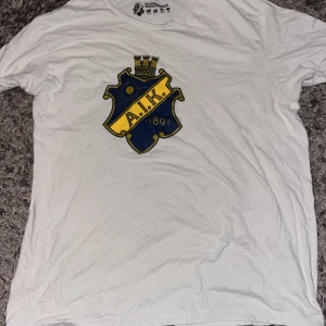 Vit AIK t-shirt med stort tryck - Vit t-shirt från AIK Official Merchandise med stort klubbmärke i blått och gult på bröstet. Klassisk rund halsringning och korta ärmar. Tillverkad i 100% bomull, perfekt för dig som vill visa ditt AIK-stöd på ett stilrent sätt.