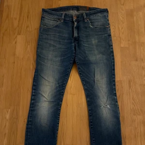 Blå Wrangler ”Larston” jeans med slitningar - Snygga blå jeans från Wrangler ”Larston” med klassisk femficksdesign och tydliga slitningar på benen. Jeansen har raka ben, normal passform och är tillverkade i robust denim. Bakfickorna har den ikoniska W-sömmen och läderpatch med logga.