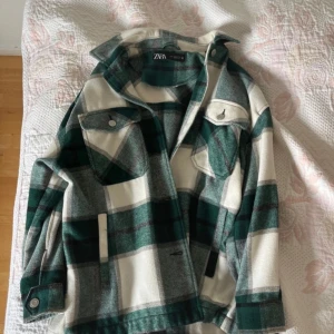 Rutig jacka från Zara XS - Grön, grå och vit rutig jacka från Zara i storlek XS. Jackan har klassisk krage, två bröstfickor med lock och knappar samt långa ärmar med knapp vid ärmslut. Perfekt lager-på-lager-plagg för säsongsskiftet.
