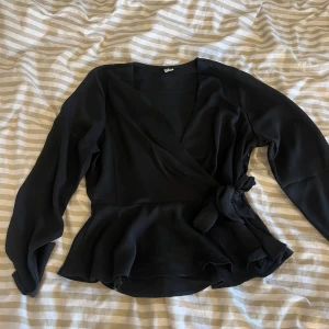 Svart omlottblus från NLY Trend - Säljer en svart omlottblus från NLY Trend i storlek 38. Blusen har långa ärmar, knyt i sidan och volangkant nedtill. Snygg v-ringning och lätt, flowigt tyg.