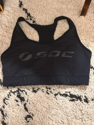 Svart sport-bh från SOC - Snygg svart sport-bh från SOC med brottarrygg och bred resår under bysten. Tillverkad i ett stretchigt funktionsmaterial som andas, perfekt för träning och aktivitet. Stor logga framtill i ton-i-ton svart.