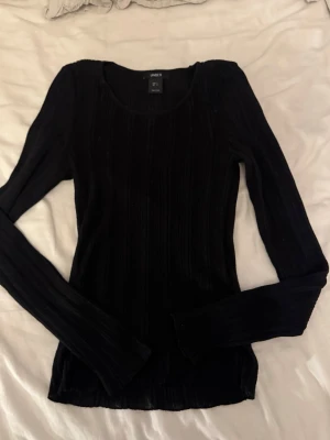 Svart ribbad tröja från Lindex - Säljer en svart långärmad tröja från Lindex med ribbad struktur. Tröjan har en rund halsringning och är figurnära i modellen. Perfekt basplagg att styla till olika outfits.