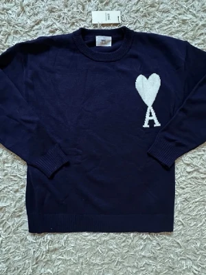 Mörkblå stickad tröja Ami Paris - Snygg mörkblå stickad tröja från Ami Paris med deras ikoniska vita hjärta och bokstaven A på bröstet. Tröjan har rund halsringning, långa ärmar och ribbade muddar. Perfekt för dig som gillar stilrena och trendiga plagg.