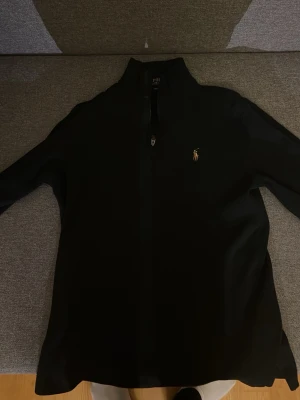 Svart ziptröja från Polo Ralph Lauren - Klassisk svart ziptröja från Polo Ralph Lauren med halv dragkedja och den ikoniska broderade loggan i gult på bröstet. Tröjan har hög krage och är tillverkad i ett mjukt ribbat material som ger en clean och stilren look.
