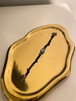 Svart armband med blått öga - Snyggt armband med svart justerbart band och en blå pärla i mitten med ögonmotiv. Perfekt accessoar för dig som gillar minimalistisk stil och vill ha något unikt på handleden. Metallspänne i silverfärg.