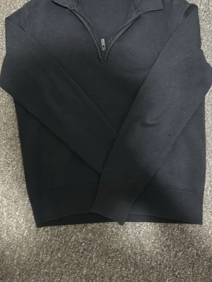 Svart halvzip-tröja från Dobber - Svart långärmad tröja från Dobber med halv dragkedja framtill och ståkrage. Enkel och stilren design, perfekt för lager-på-lager. Mjuk bomullskänsla och ribbade muddar vid ärmslut och nederkant.