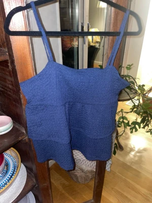 Blå linnetopp med volang - Snygg blå linnetopp med smala axelband och volangdetaljer. Toppen har en strukturerad yta och är perfekt för varma dagar. Enkel att matcha med både jeans och kjol.