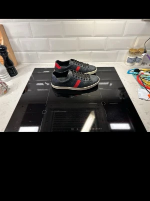 Gucci ace skor  - Snygga svarta sneakers med röd och mörkblå rand på sidan samt vit sula. Skorna har lågt skaft och klassisk snörning. Materialet ser ut att vara läder eller mocka. Perfekta för en stilren och sportig look.