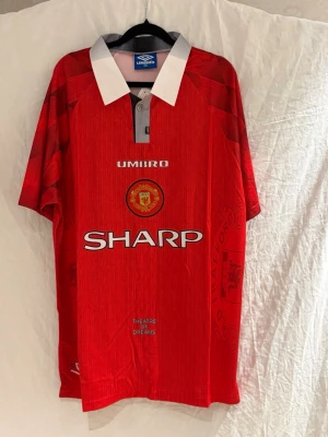 Manchester United cantona #7 - säljer en retro Manchester United tröja från i rött med vita detaljer och klubbmärke på bröstet. SHARP-logga på magen och texten 'THEATRE OF DREAMS' nedtill. Kortärmad med krage och knappar. Perfekt för fotbollsfans!