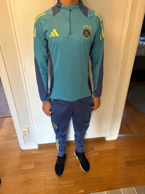 Ajax träningsset blå/gul Adidas - Ajax träningsset från Adidas i blå och turkos med gula detaljer. Setet består av långärmad tröja med halv dragkedja och matchande byxor, båda med Ajax-logga och Adidas ränder. Perfekt för fotbollsträning eller sportiga dagar. Personen i bilden är 180 cm 