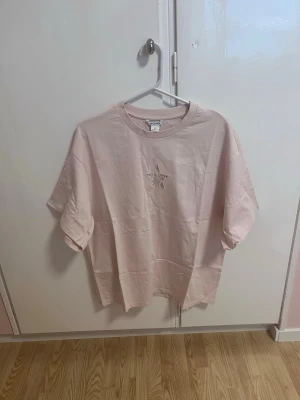 Oversized T-shirt  - Snygg ljusrosa t-shirt från Monki med en broderad stjärna på bröstet. T-shirten har en klassisk oversized passform och rund halsringning. Perfekt för en avslappnad och trendig look.