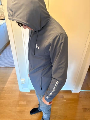 Grå vindjacka från Under Armour - Snygg grå vindjacka från Under Armour med huva och dragkedja. Jackan har logga på bröstet och text på ärmen, samt praktiska fickor framtill. Perfekt för blåsiga dagar och sportiga outfits. Personen längd i bilden är 180cm