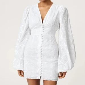 Adoore FLORENCE DRESS  - Supersnygg vit kort klänning från Adoore med v-ringning och puffiga långärmar. Klänningen har broderade blommor, knappar framtill och markerad midja som ger en slim passform. Perfekt för sommarens alla tillfällen, med romantiska detaljer och lätt tyg. Storlek 36.