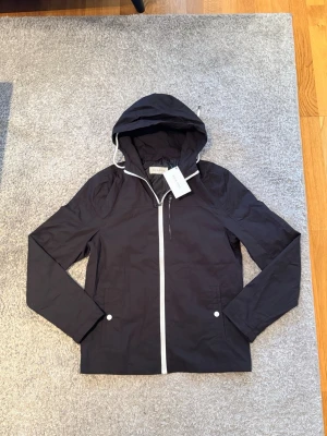 Svart vindjacka från Pier One storlek S - Stilren svart vindjacka från Pier One med vit dragkedja och vita detaljer vid fickorna. Jackan har huva med dragsko och är tillverkad i ett lätt, vindtätt material. Perfekt för blåsiga dagar och enkel att matcha med olika outfits. Storlek S