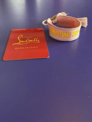 Christian Dior tygarmband rosa/gul - Säljer ett snyggt tygarmband från Christian Dior i ljusrosa och gult med broderad text. Armbandet har justerbar knytning med tofsar i ändarna och ger en trendig och avslappnad vibe. Perfekt accessoar för att lyfta din outfit.