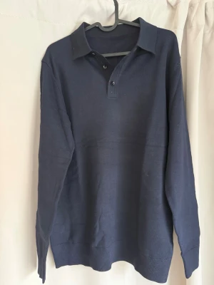 Mörkblå pikétröja med lång ärm från nuvéclothing - Klassisk mörkblå pikétröja med lång ärm och krage. Tröjan har knäppning med tre knappar vid halsen och ribbade muddar vid ärmslut och nederkant. Perfekt för en stilren och enkel look.