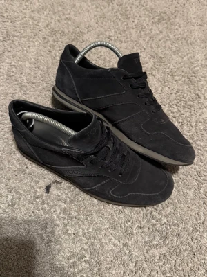 Svarta sneakers från TOUS i mocka - Stilrena svarta sneakers från TOUS i mjuk mocka med matchande svarta skosnören och diskret logga på sidan. Skorna har en låg silhuett och en grå sula som ger en clean look. Perfekta för dig som gillar minimalistisk och tidlös stil.