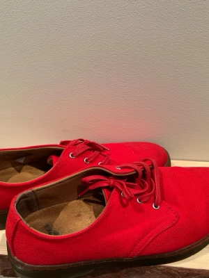 Röda loafers från Dr. Martens - Säljer ett par röda loafers från Dr. Martens i tyg med klassisk snörning och låga sulor. Skorna har en enkel och stilren design med metallöljetter och bruna innersulor. Perfekta för dig som vill sticka ut med färg på fötterna.