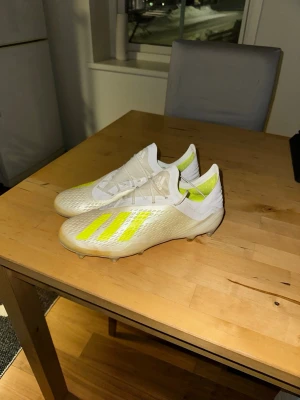 Adidas vita och gula fotbollsskor - Säljer ett par snygga Adidas fotbollsskor i vitt med gula detaljer och klassiska tre ränder. Skorna har låg profil, snörning och ovandel i syntetmaterial med textur för bättre bollkontroll. Perfekta för dig som vill sticka ut på planen.