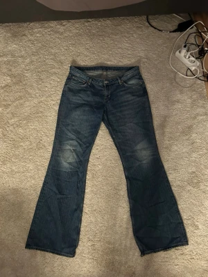 Blå bootcut jeans med slitningar - Säljer ett par klassiska blå bootcut jeans med lätt slitna detaljer vid knäna. Jeansen har fem fickor, normalhög midja och utsvängda ben. Perfekta för en avslappnad och trendig look. Dom är lågmidjade 