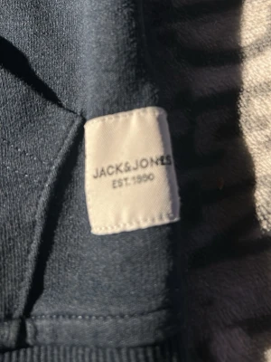 Blå hoodie från Jack & Jones - Snygg blå hoodie från Jack & Jones med dragkedja framtill och två fickor. Tillverkad i mjukt bomullsmaterial och har ribbade muddar vid ärmslut och nederkant. Perfekt för en avslappnad och stilren look.