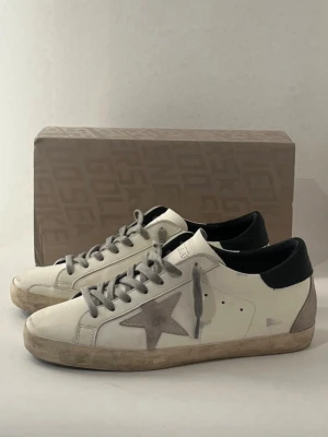Golden Goose Super-Star vita sneakers - Golden Goose Super-Star sneakers i vitt läder med grå stjärndetalj på sidan, grå skosnören och svart hälparti. Klassisk låg modell med slitna detaljer för en trendig look. Snygg kontrast mellan färgerna och ikonisk branding på insidan.