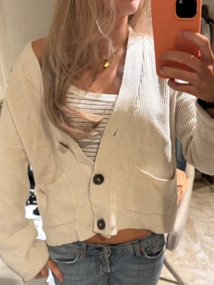 Vit ribbad kofta med stora knappar - Säljer en krämvit ribbad kofta med stora mörka knappar och två fickor framtill. Koftan har en avslappnad, lite boxig passform och är gjord i ett mjukt stickat material. Perfekt att slänga över ett linne eller t-shirt för en chill look.