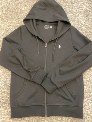 Polo Ralph Lauren zip - Snygg svart hoodie från Polo Ralph Lauren med dragkedja framtill, två fickor och klassisk vit logga på bröstet. Tillverkad i mjukt bomullsmaterial och har justerbar huva med snören. Perfekt för en chill och stilren look.