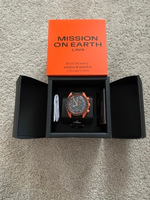 Omega x Swatch MoonSwatch Lava - Säljer en Omega x Swatch Speedmaster MoonSwatch Mission on Earth Lava. Klockan har svart urtavla med orange detaljer och boett i biokeramik. Svart textilarmband med vita och orange sömmar samt loggor. Kommer i originalförpackning.