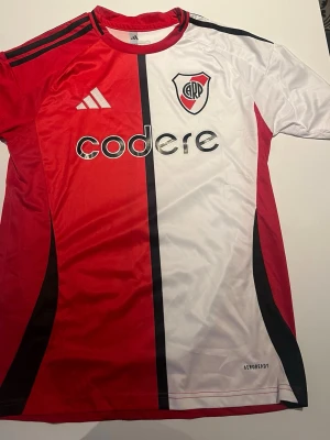 River Plate matchtröja Adidas röd/vit - River Plate matchtröja från Adidas i rött och vitt med svart detalj. Tröjan har klubbmärke, sponsorloggor och Aeroready-material som andas. Perfekt för fotbollsträning eller att bära på läktaren.