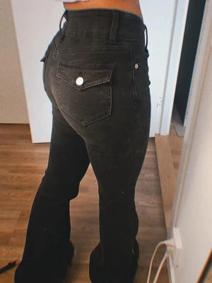 Svarta bootcut jeans med mid/low waist - Snygga svarta jeans med bootcut-modell och mid waist. Jeansen har klassiska fickor bak med silverfärgade knappar och är tillverkade i stretchigt jeansmaterial. Perfekta för dig som gillar en trendig och bekväm passform. Nya men jag föredrar högre jeans, storlek XS ungefär 25/26 i jeans.