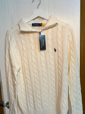 Vit kabelstickad tröja Polo Ralph Lauren - Kabelstickad vit tröja från Polo Ralph Lauren med halv dragkedja och broderad logga på bröstet. Tröjan har ribbade muddar och krage, perfekt för en clean och klassisk look. Tillverkad i mjukt material som känns lyxigt. Perfekt för kylan som sveper över landet nu!